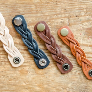 K&ouml;nnte beinhalten: Vier geflochtene Lederarmb&auml;nder mit Metallschlie&szlig;en. Die Armb&auml;nder sind in verschiedenen Farben: beige, schwarz, braun und orange.