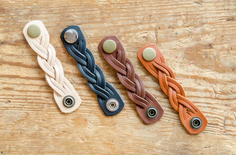 K&ouml;nnte beinhalten: Vier geflochtene Lederarmb&auml;nder mit Metallschlie&szlig;en. Die Armb&auml;nder sind in verschiedenen Farben: beige, schwarz, braun und orange.