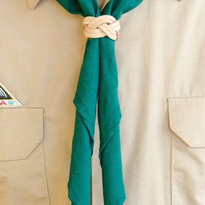 K&ouml;nnte beinhalten: Ein gr&uuml;nes Halstuch, das mit einem Ledergleiter verknotet ist. Das Halstuch wird auf einem braunen Uniformhemd getragen.