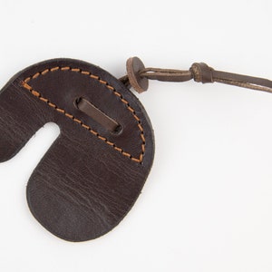 Può includere: Un puntale per dita da tiro con l'arco in pelle marrone con un cinturino in pelle marrone e dettagli di cucitura marrone.