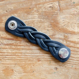 K&ouml;nnte beinhalten: Ein schwarzes Lederarmband mit geflochtenem Design und silbernen Metall-Druckkn&ouml;pfen.