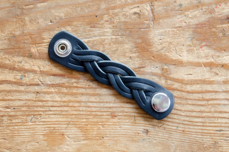 K&ouml;nnte beinhalten: Ein schwarzes Lederarmband mit geflochtenem Design und silbernen Metall-Druckkn&ouml;pfen.