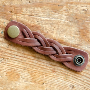 K&ouml;nnte beinhalten: Ein braunes Lederarmband mit geflochtenem Design und zwei Metallschlie&szlig;en. Die Schlie&szlig;en sind rund und haben eine gl&auml;nzende Oberfl&auml;che.