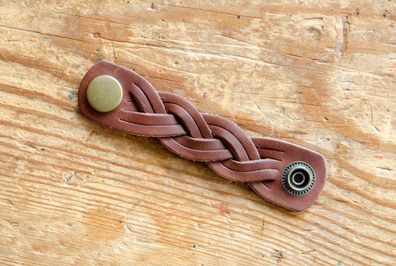 K&ouml;nnte beinhalten: Ein braunes Lederarmband mit geflochtenem Design und zwei Metallschlie&szlig;en. Die Schlie&szlig;en sind rund und haben eine gl&auml;nzende Oberfl&auml;che.
