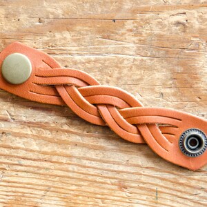 K&ouml;nnte beinhalten: Ein geflochtenes Lederarmband in Braun mit zwei Metallverschl&uuml;ssen. Das Armband ist in einem einfachen Flechtmuster gewebt.