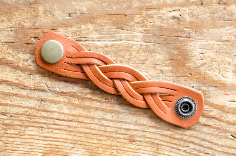 K&ouml;nnte beinhalten: Ein geflochtenes Lederarmband in Braun mit zwei Metallverschl&uuml;ssen. Das Armband ist in einem einfachen Flechtmuster gewebt.