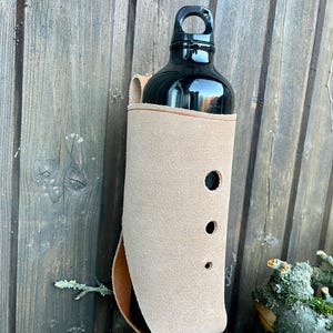 Peut inclure: Une gourde noire dans un étui en cuir beige avec trois découpes circulaires. Le support est fixé à une surface en bois. La bouteille a un bouchon noir avec une boucle de transport.