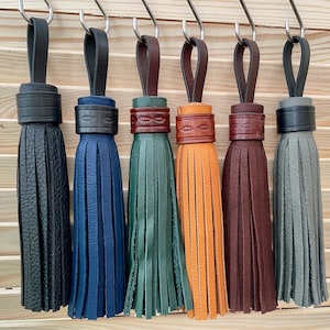Op de afbeelding: Een verzameling leren kwasten in verschillende kleuren, waaronder zwart, blauw, groen, oranje, bruin en grijs. Elke kwast hangt aan een metalen haak en heeft een lus aan de bovenkant en een decoratieve band.