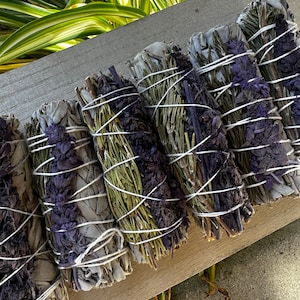 May include: A bundle of sage smudge sticks with lavender and rosemary. The sticks are tied together with white string and are arranged on a wooden surface.