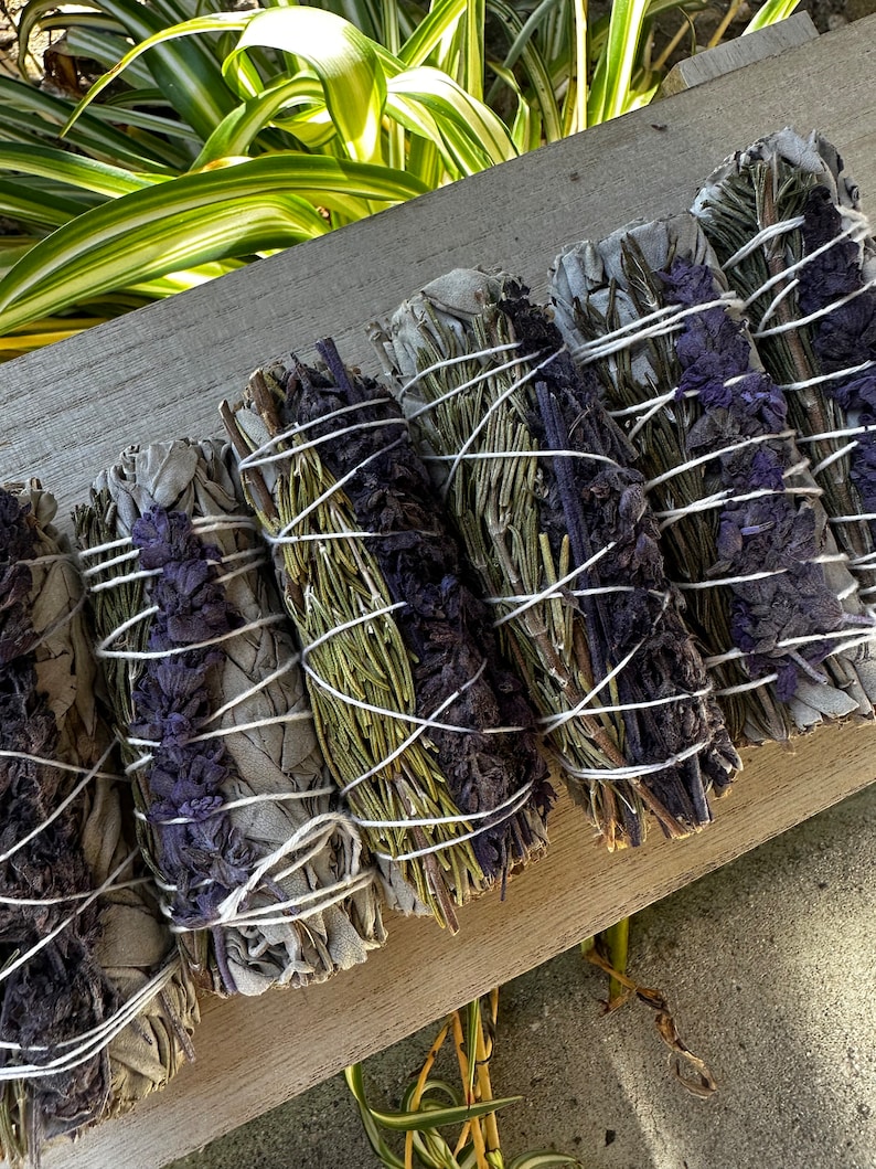 May include: A bundle of sage smudge sticks with lavender and rosemary. The sticks are tied together with white string and are arranged on a wooden surface.