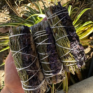 May include: Three bundles of smudge sticks, tied with white string. The bundles are made of dried herbs, including sage, lavender, and rosemary.