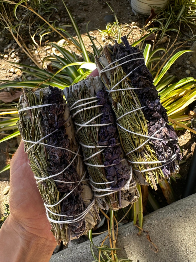 May include: Three bundles of smudge sticks, tied with white string. The bundles are made of dried herbs, including sage, lavender, and rosemary.