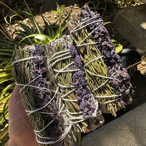 May include: Three bundles of smudge sticks, tied with twine, containing dried herbs and flowers. The bundles are a mix of green, white, and purple.