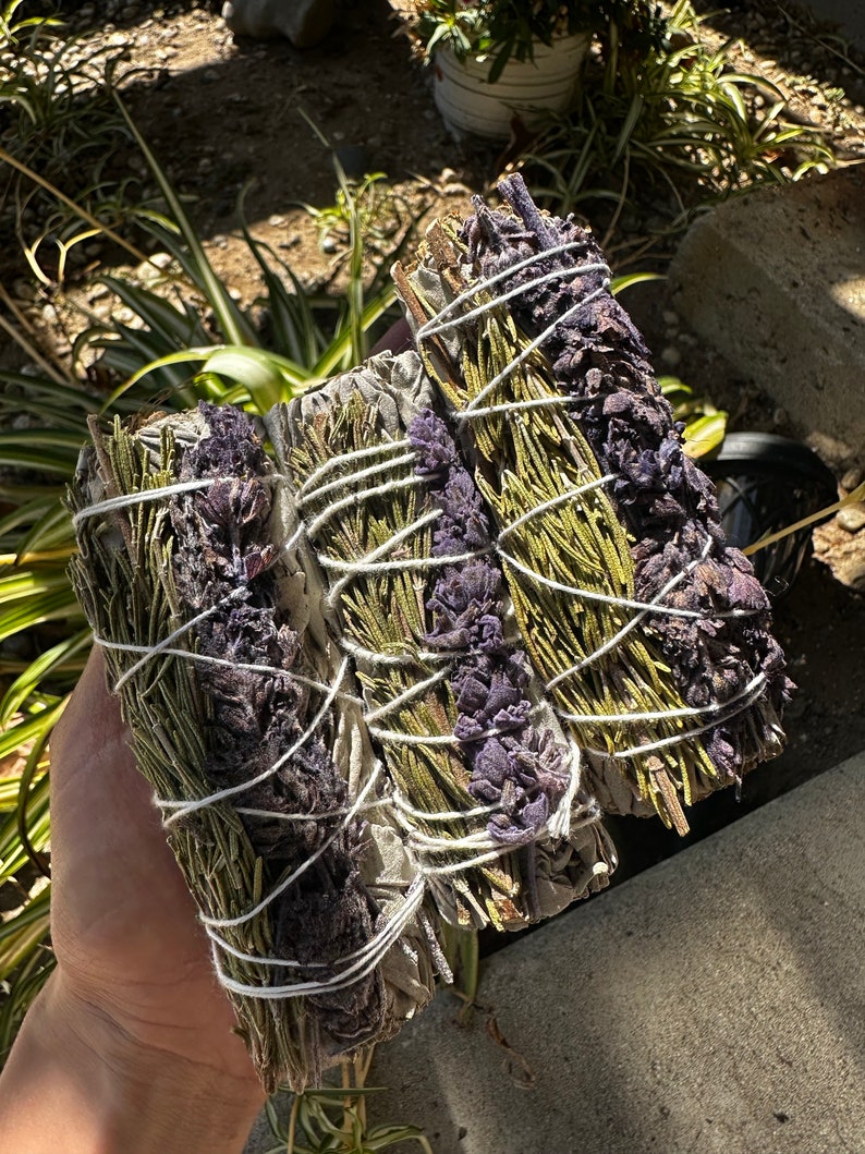 May include: Three bundles of smudge sticks, tied with twine, containing dried herbs and flowers. The bundles are a mix of green, white, and purple.