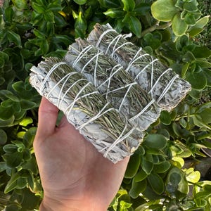 May include: Three bundles of white sage and rosemary smudge sticks tied together with twine. The smudge sticks are a light green color and are wrapped in white paper.