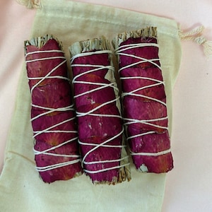 May include: Three bundles of white sage smudge sticks wrapped with dried rose petals and white twine. The bundles are arranged on a white cloth background.