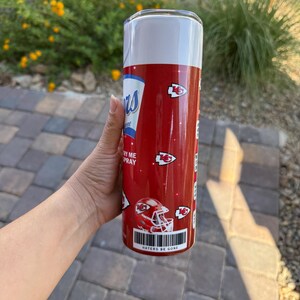 May include: A red and white tumbler with a design resembling a spray can. The tumbler features the text "Chiefs" and "Haters Be Gone", along with images of football helmets and team logos. The tumbler is cylindrical and appears to be made of metal.