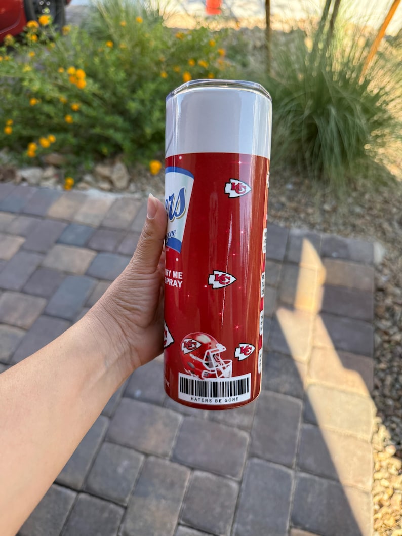 May include: A red and white tumbler with a design resembling a spray can. The tumbler features the text "Chiefs" and "Haters Be Gone", along with images of football helmets and team logos. The tumbler is cylindrical and appears to be made of metal.