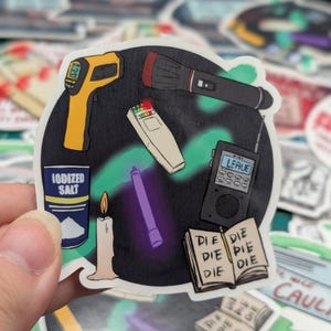 May include: A sticker featuring a collection of ghost hunting tools, including a flashlight, a thermometer, a candle, a spirit box, a UV light, and a container of iodized salt. The sticker also includes a notebook with the word "DIE" written on each page.
