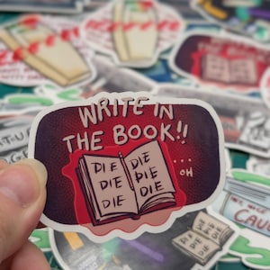 May include: A sticker with a red book that says "WRITE IN THE BOOK!!" and has the word "DIE" written on each page. The book is open and there is a pencil on the top page. The sticker is surrounded by other stickers.