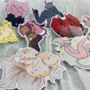 May include: A set of nine colorful stickers featuring various characters and designs. The stickers include a cat, a fish, a lion, a demon, a person with a halo, a person with a fish tail, and a person with wings. The stickers are all on a light green background.