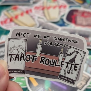 May include: A sticker with the text "Meet me at Tanglewood for some Tarot Roulette". The sticker features a cartoon illustration of a tarot reading with candles and three tarot cards: High Priestess, Hanged Man, and a card with a mountain scene.