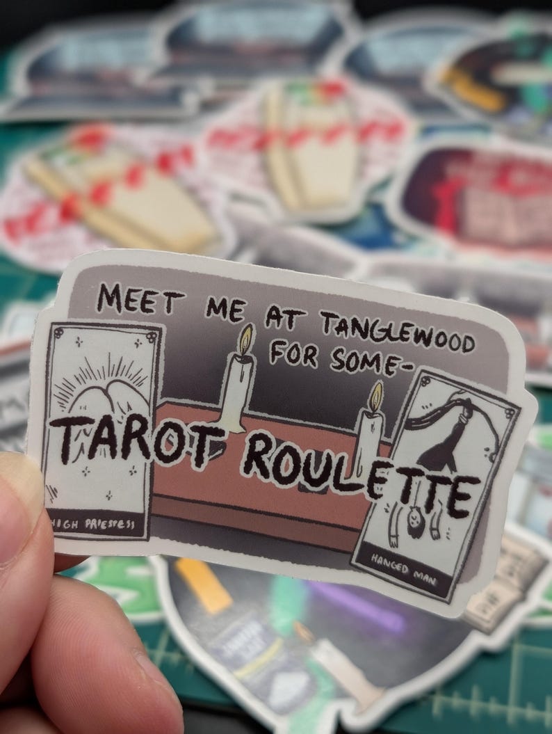 May include: A sticker with the text "Meet me at Tanglewood for some Tarot Roulette". The sticker features a cartoon illustration of a tarot reading with candles and three tarot cards: High Priestess, Hanged Man, and a card with a mountain scene.