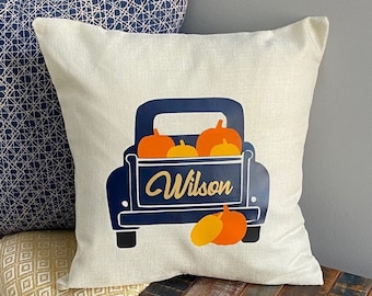 Könnte beinhalten: Ein weißes Kissen mit einem blauen Lastwagen, der orangefarbene Kürbisse transportiert. Der Lastwagen hat den Schriftzug "Wilson" auf der Rückseite. Das Kissen liegt auf einer Holzbank mit einer gelben und weißen Musterdecke.