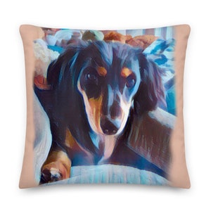 May include: A close-up of a black and tan dachshund with brown eyes looking directly at the camera. The dog is lying on a blue and white patterned pillow with a peach-colored background.