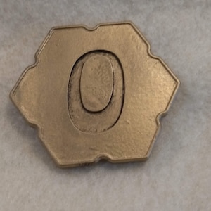 May include: A gold-colored metal token with a hexagonal shape and the number "0" embossed on the front.