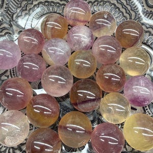 May include: A collection of polished, spherical gemstones in shades of purple, yellow, and brown. The spheres are arranged in a decorative, silver-colored dish with a patterned rim. The gemstones have a translucent quality, with internal color variations.