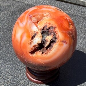 May include: A polished carnelian sphere featuring swirling bands of orange and red hues, with a prominent, textured, dark brown and white crystalline geode cavity exposed on its surface, resting on a small, circular, dark brown wooden stand.