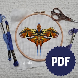 May include: A cross-stitch project featuring a geometric design of a winged creature in shades of orange, brown, and black, set within a wooden embroidery hoop. Blue embroidery floss and scissors are also visible. The image includes the text "PDF".