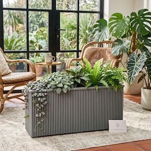 Op de afbeelding: Een rechthoekige grijze plantenbak met een verticale geribbelde textuur, gevuld met diverse groene kamerplanten, waaronder een hangplant. Een klein wit kaartje met de tekst Etsy.com/TheBotanicalRoom ligt op de vloer naast de bak.