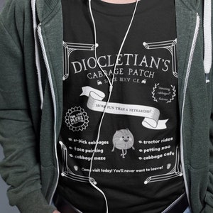 May include: A person wearing a green hooded sweatshirt over a black t-shirt with a white graphic design that says "Diocletian's Cabbage Patch" with a cartoon cabbage character. The text also says "More fun than a Tetrarchy!" and lists activities like "u-pick cabbages", "face painting", "tractor rides", "petting zoo", and "cabbage cafe".