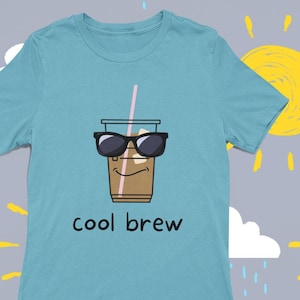 May include: A light blue t-shirt featuring a cartoon illustration of a cup of iced coffee wearing sunglasses and a straw. The text "cool brew" is printed below the image.