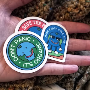 May include: Three round stickers with a green, blue, and red background. The stickers feature a cartoon earth with a smiling face. The text on the stickers reads "Don't Panic It's Organic", "Save The Planet", and "Make Earth Cool Again".