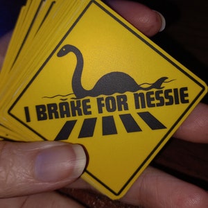Puede incluir: Una pila de señales de advertencia amarillas con una silueta negra del monstruo del Lago Ness y el texto "I Brake For Nessie".