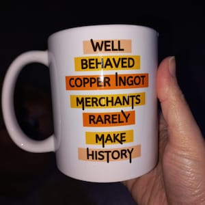 May include: White ceramic mug with a curved handle. The mug features the text "WELL BEHAVED COPPER INGOT MERCHANTS RARELY MAKE HISTORY" in a mix of orange, yellow, and black fonts. The mug is held against a dark background.