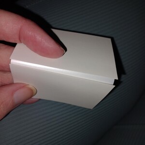 May include: A small, white, rectangular object with a folded design. The object is held by a person's fingers, with the focus on the clean lines and the way the light reflects off the surface. The object appears to be made of paper or cardstock.