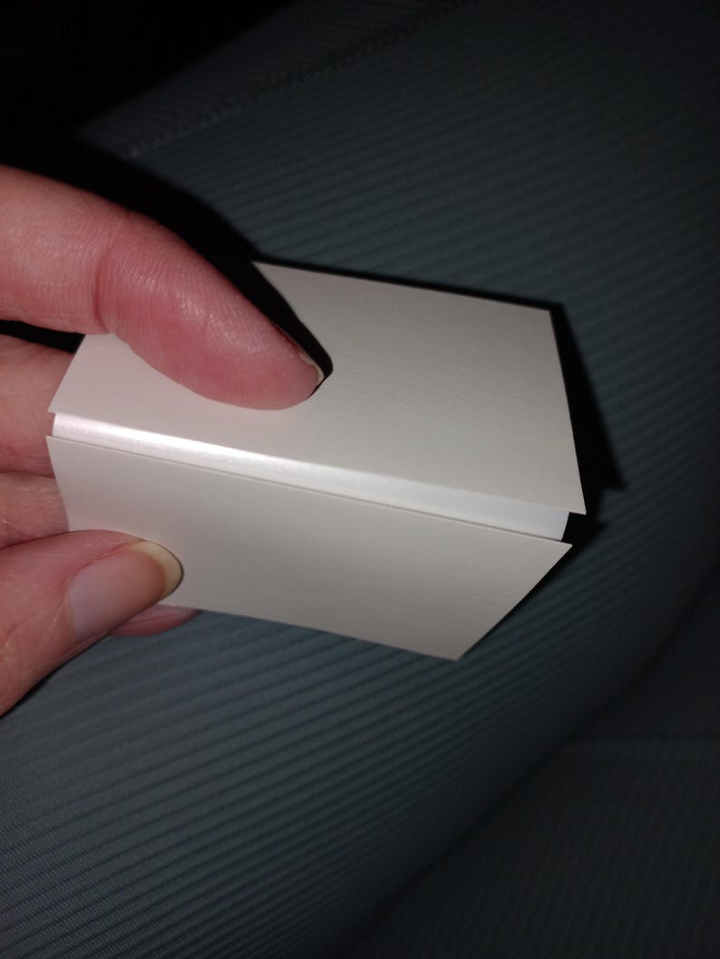 May include: A small, white, rectangular object with a folded design. The object is held by a person's fingers, with the focus on the clean lines and the way the light reflects off the surface. The object appears to be made of paper or cardstock.