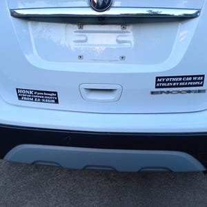 Może przedstawiać: Biały samoch&oacute;d z dwoma czarnymi naklejkami na zderzaku. G&oacute;rna naklejka głosi "MY OTHER CAR WAS STOLEN BY SEA PEOPLE", a dolna naklejka głosi "HONK if you bought SURPAR COPPER INGOTS FROM EA-NASIR".