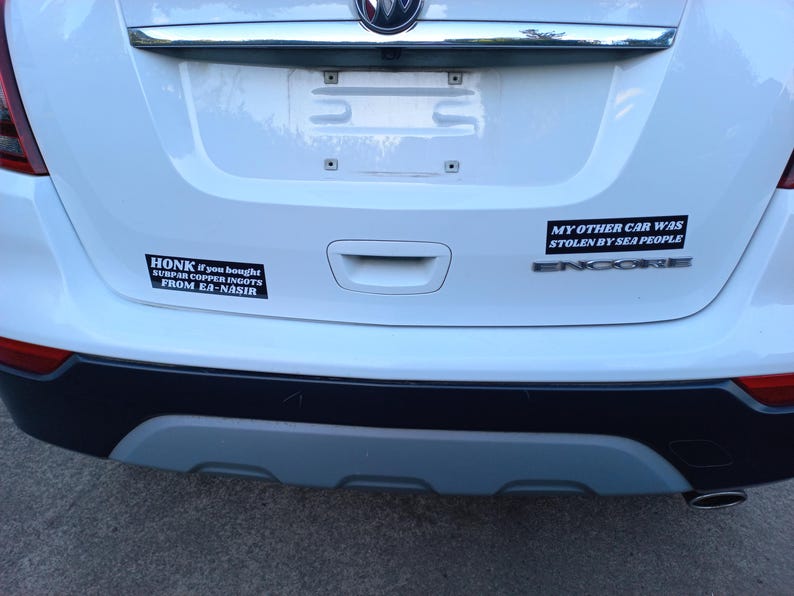 Może przedstawiać: Biały samoch&oacute;d z dwoma czarnymi naklejkami na zderzaku. G&oacute;rna naklejka głosi "MY OTHER CAR WAS STOLEN BY SEA PEOPLE", a dolna naklejka głosi "HONK if you bought SURPAR COPPER INGOTS FROM EA-NASIR".