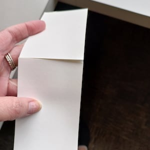 May include: A stack of blank, white rectangular cards held near a white box. The cards are smooth and have a slight curve. The background is a dark brown surface and a white box.