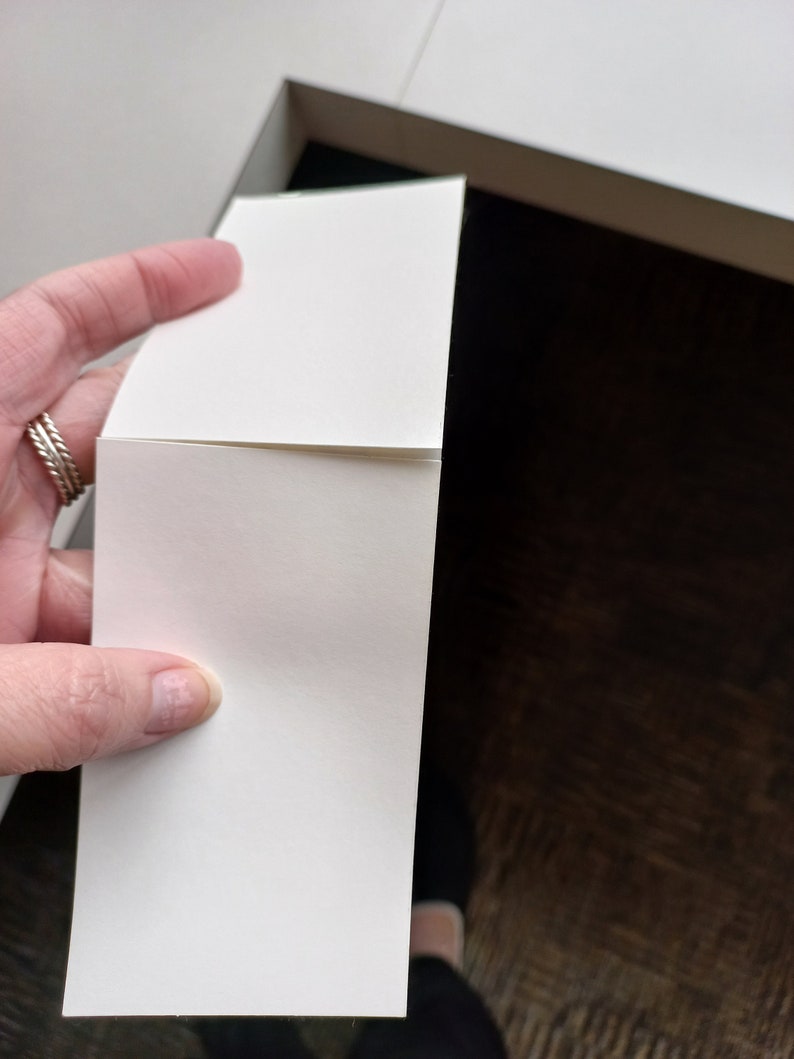May include: A stack of blank, white rectangular cards held near a white box. The cards are smooth and have a slight curve. The background is a dark brown surface and a white box.