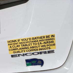 May include: A yellow bumper sticker with black text that reads "HONK IF YOU'D RATHER BE IN ANCIENT MESOPOTAMIA INSCRIBING A CLAY TABLET TO EA-NĀṢIR, COMPLAINING ABOUT HIS POOR QUALITY COPPER INGOTS." Below the sticker is a silver "ENCORE" emblem and a blue and green emblem.