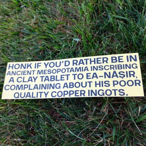 May include: A yellow rectangular sign with black text reading "HONK IF YOU'D RATHER BE IN ANCIENT MESOPOTAMIA INSCRIBING A CLAY TABLET TO EA-NĂŞIR, COMPLAINING ABOUT HIS POOR QUALITY COPPER INGOTS." The sign is on a bed of green grass.