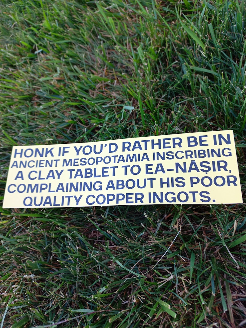 May include: A yellow rectangular sign with black text reading "HONK IF YOU'D RATHER BE IN ANCIENT MESOPOTAMIA INSCRIBING A CLAY TABLET TO EA-NĂŞIR, COMPLAINING ABOUT HIS POOR QUALITY COPPER INGOTS." The sign is on a bed of green grass.