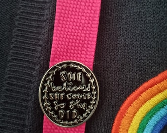 May include: A black and silver pin with the text "She believed she could so she did" on a black sweater with a rainbow embroidered patch.