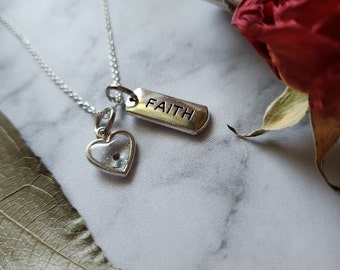 May include: A silver necklace with two charms. The first charm is a small heart with a clear glass centre. The second charm is a rectangular tag with the word "FAITH" engraved on it.